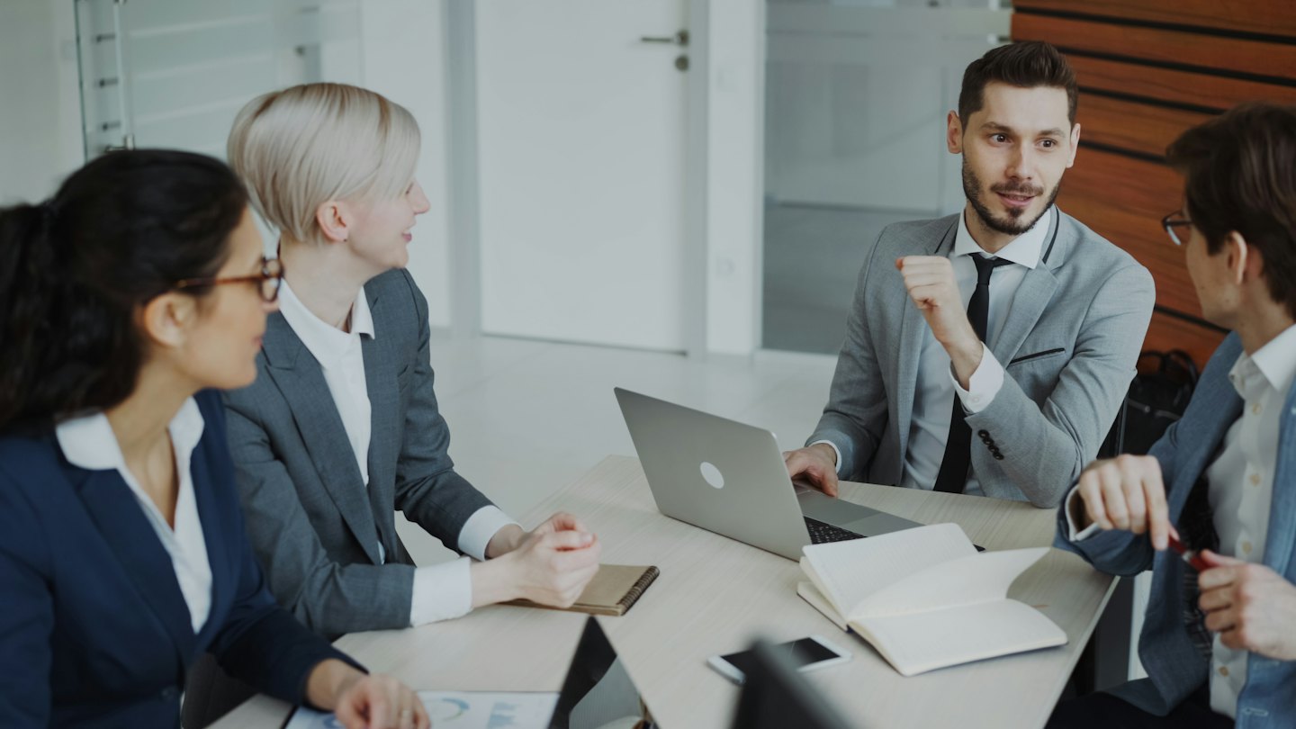 Professionals discuss business around a conference table with laptops and documents.